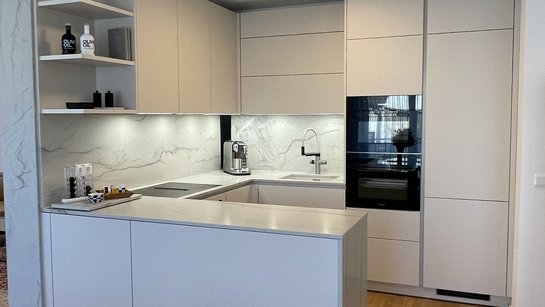Helle U-Küche mit grifflosen Fronten in angora, Natursteinarbeitsplatte und angrenzender beleuchteter Glasvitrine.