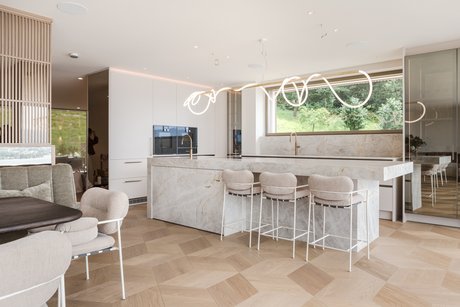 Moderne, offene Wohnküche mit großzügiger Kücheninsel mit Steinarbeitsplatte, integriertem Barbereich mit gepolsterten Hockern, sandfarbenen Fronten und Messingarmaturen sowie großflächiger Fensterfront mit Blick ins Grüne.