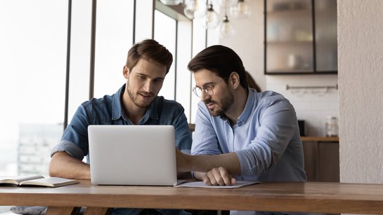 Zwei junge Männer sitzen an einem Tisch vor einem Laptop